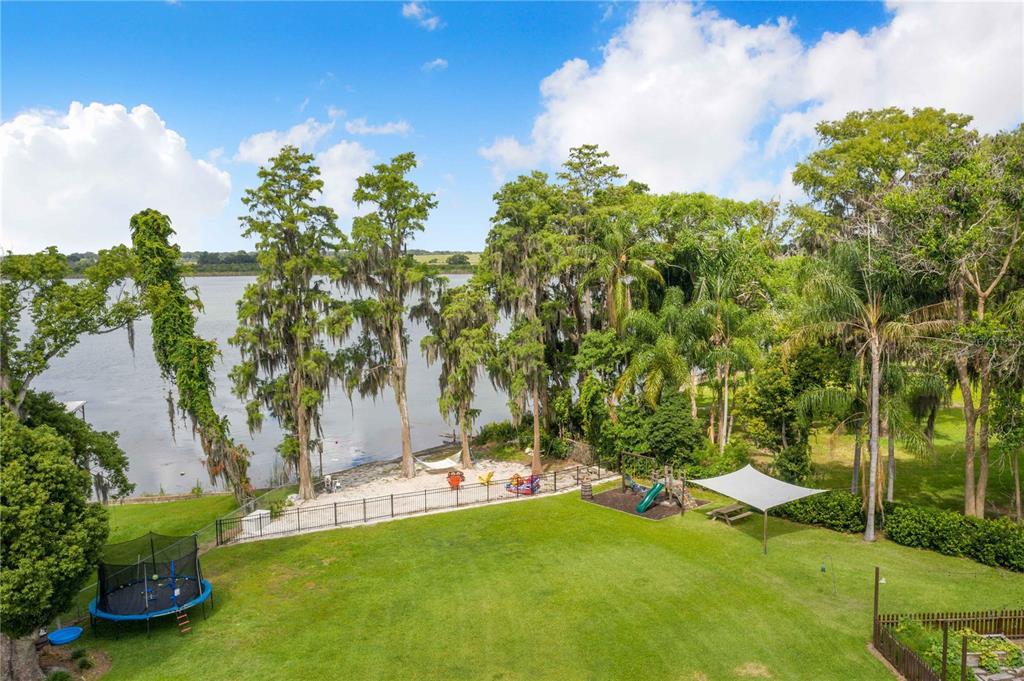 An aerial view of a grassy backyard featuring a trampoline, playground, and waterfront beach area — 775 S BUENA VISTA DRIVE, LAKE ALFRED, FL
