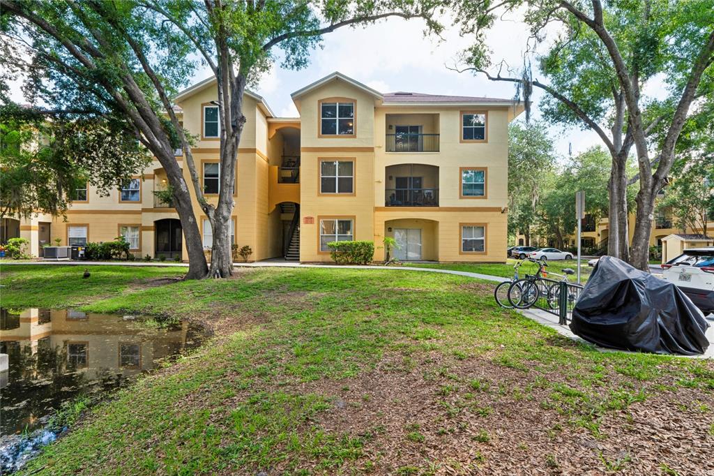 A three-story apartment building with yellow stucco walls stands beside a small pond — 5618 PINNACLE HEIGHTS CIRCLE UNIT 312, TAMPA, FL