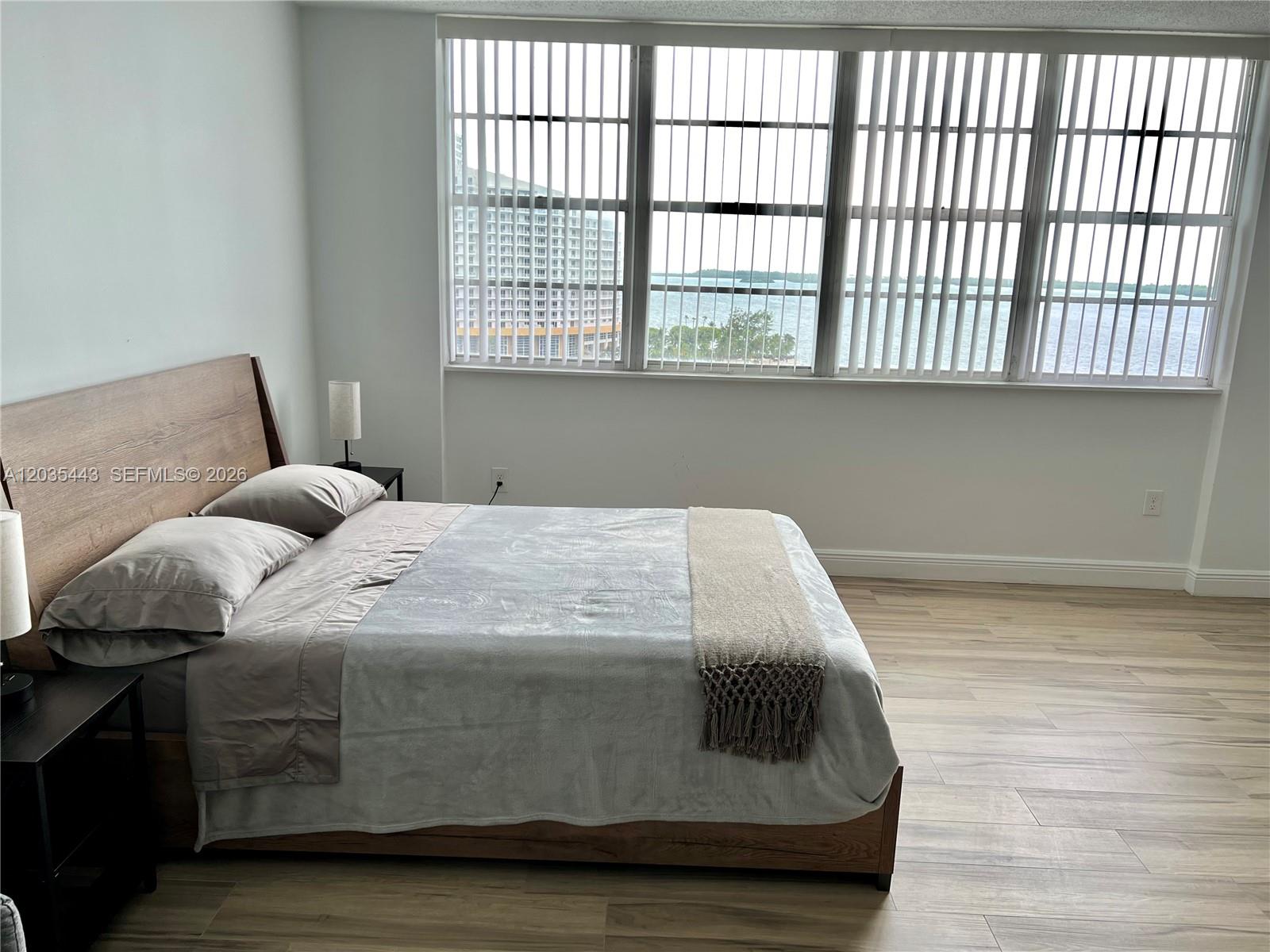 A bedroom features a wooden bed frame, light wood-look flooring, and large windows overlooking water — 801 Brickell Bay Dr # 1469, Miami FL 33131, MIAMI, FL