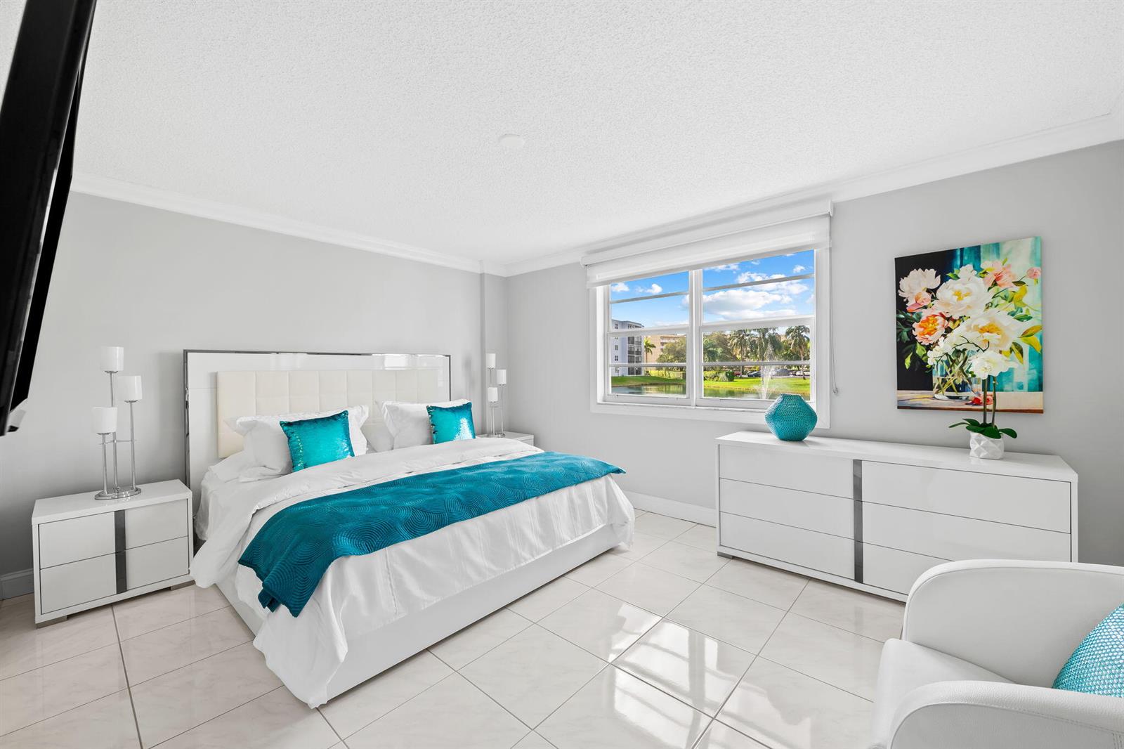 A bedroom features a white bed with teal accents, a dresser, and a window view — 170 SE 5th Avenue # 103, Dania Beach FL 33004, DANIA BEACH, FL