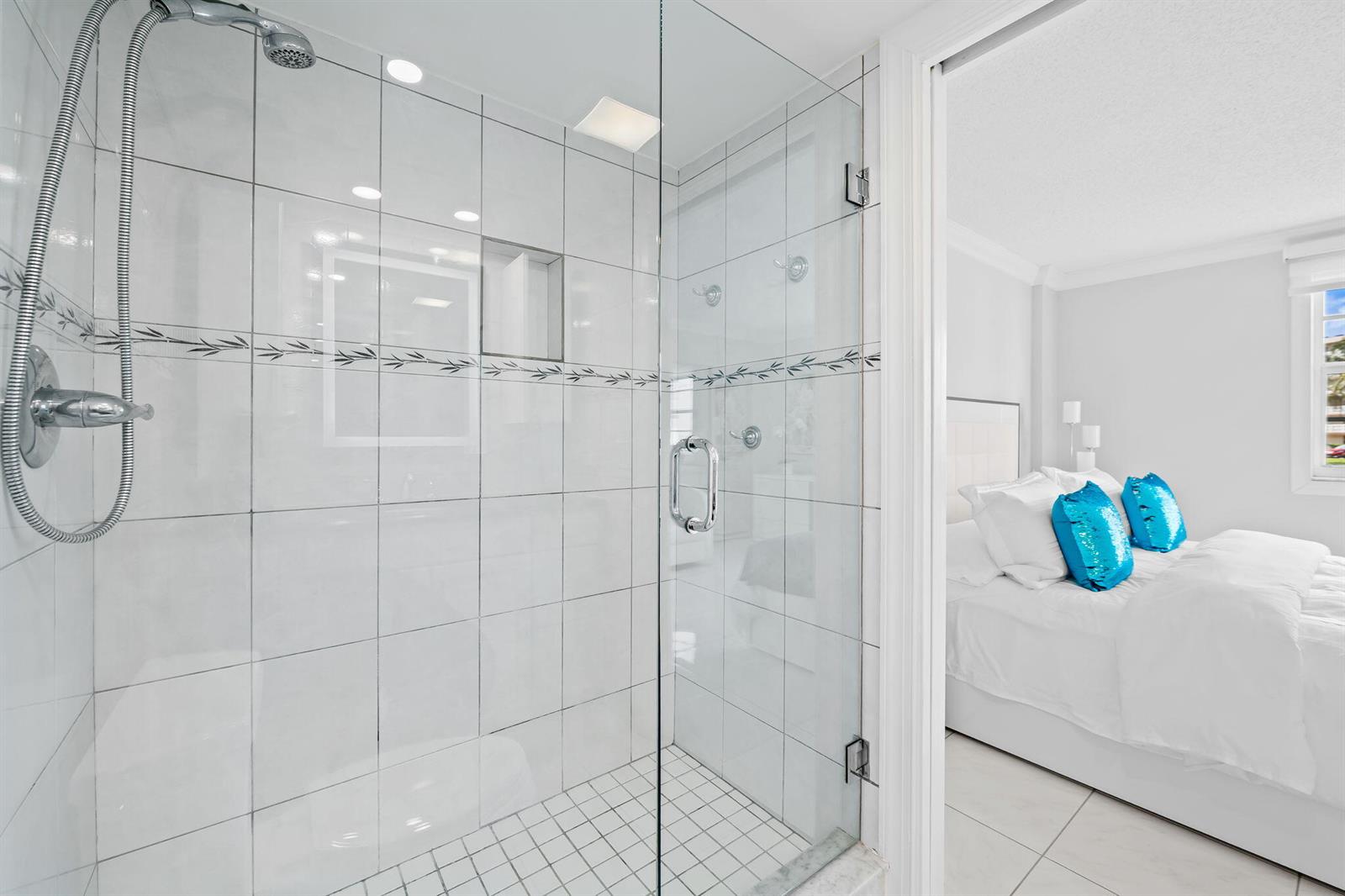 A glass-enclosed shower with white tiled walls sits adjacent to a bedroom with white bedding — 170 SE 5th Avenue # 103, Dania Beach FL 33004, DANIA BEACH, FL