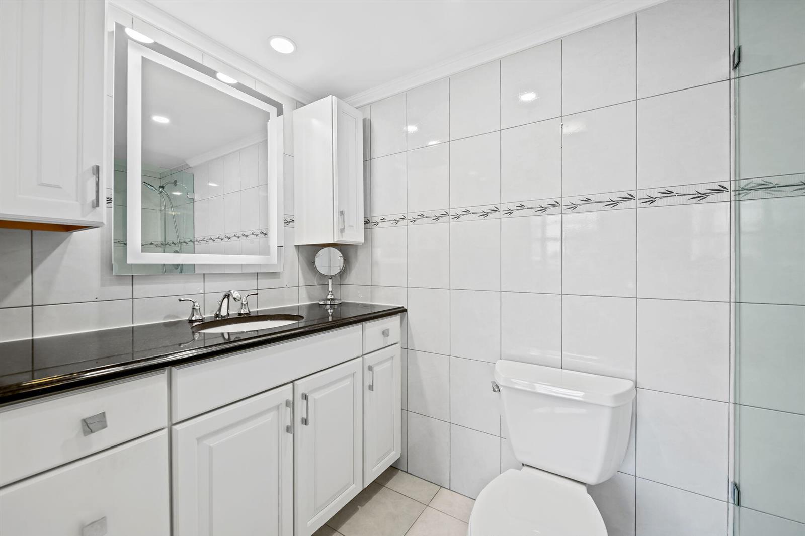 A bathroom features white cabinetry, a black countertop, a vanity mirror, and white wall tiles — 170 SE 5th Avenue # 103, Dania Beach FL 33004, DANIA BEACH, FL