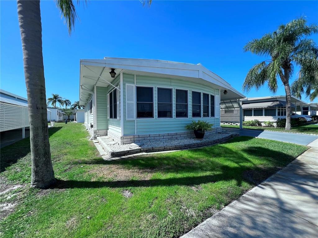 A light green manufactured home with a brick base and a carport under a blue sky — 1100 BELCHER ROAD S UNIT 772, LARGO, FL