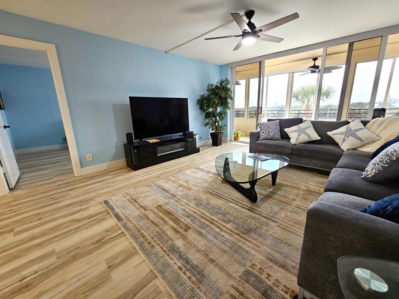 A living room features light blue walls, wood-look flooring, a gray sectional, and a balcony — 1357 NE Ocean Boulevard # 312, Stuart FL 34996, STUART, FL