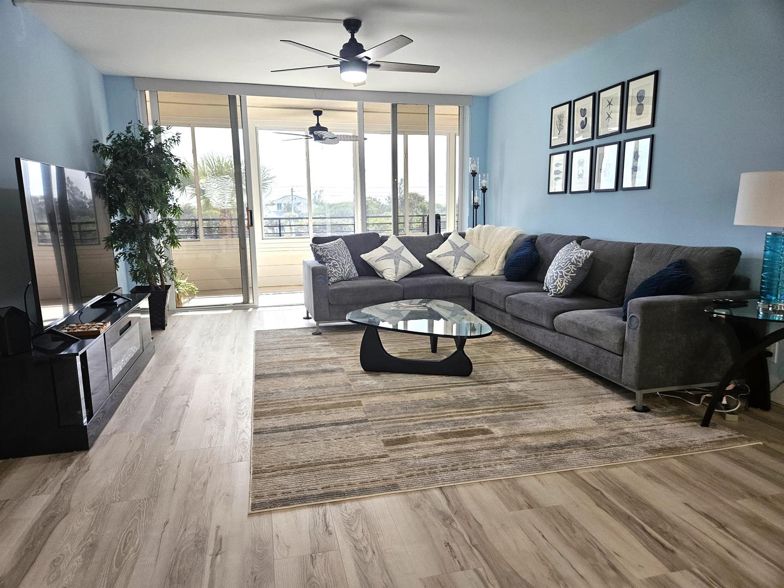 A living room features a grey sectional sofa, glass coffee table, and light wood flooring — 1357 NE Ocean Boulevard # 312, Stuart FL 34996, STUART, FL