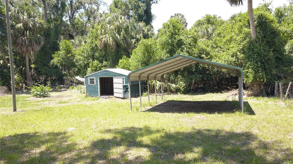 A green storage shed and a metal carport stand in a grassy, wooded backyard — 11291 SE 202ND LANE, INGLIS, FL