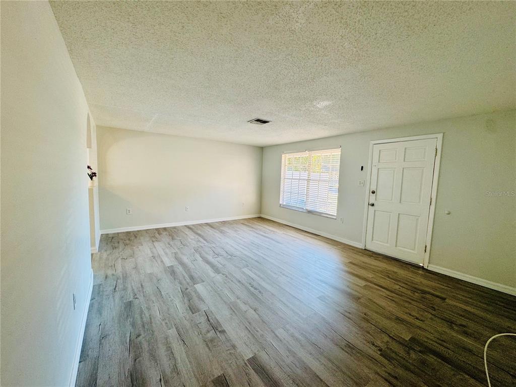 An empty living room features light gray walls, wood-look flooring, and a white front door — 7116-7118 GREEN STREET, NEW PORT RICHEY, FL