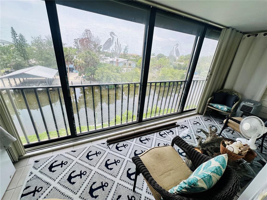 A sunroom features an anchor-patterned rug, wicker furniture, and large windows overlooking a canal — 4212 126TH STREET W UNIT 407, CORTEZ, FL