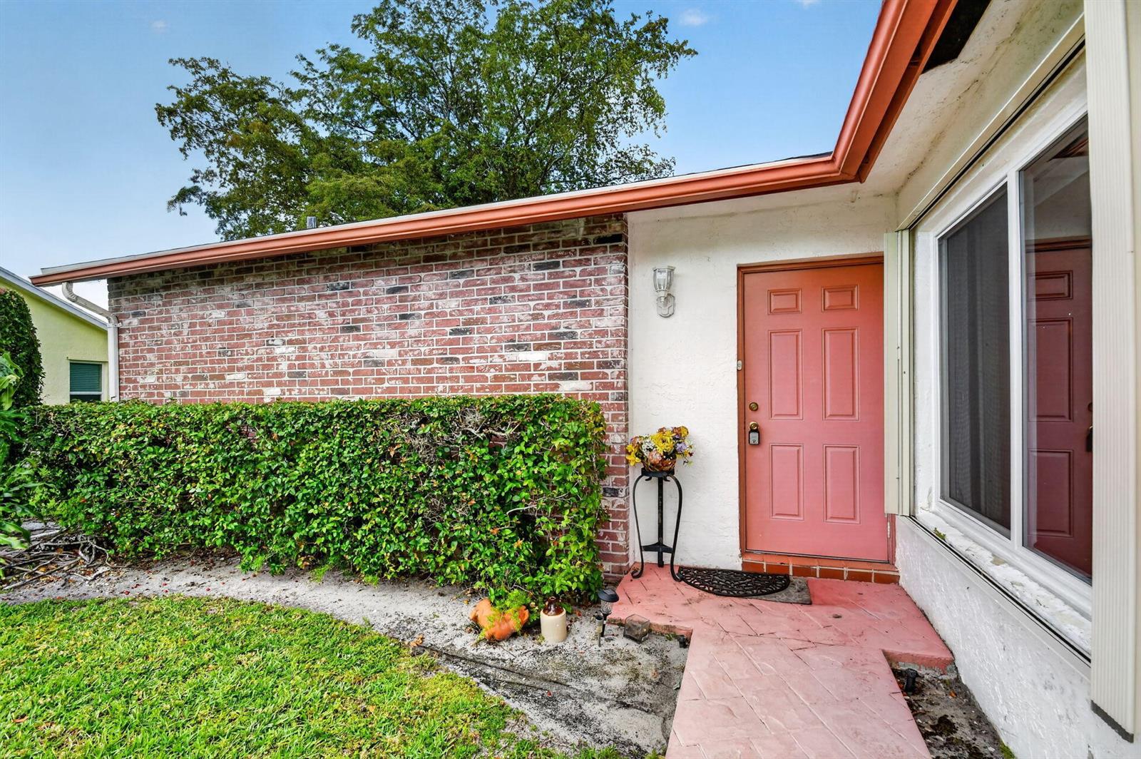 A pink front door sits beside a brick wall and a manicured hedge — 6312 Summer Sky Lane, FL 33463, FL