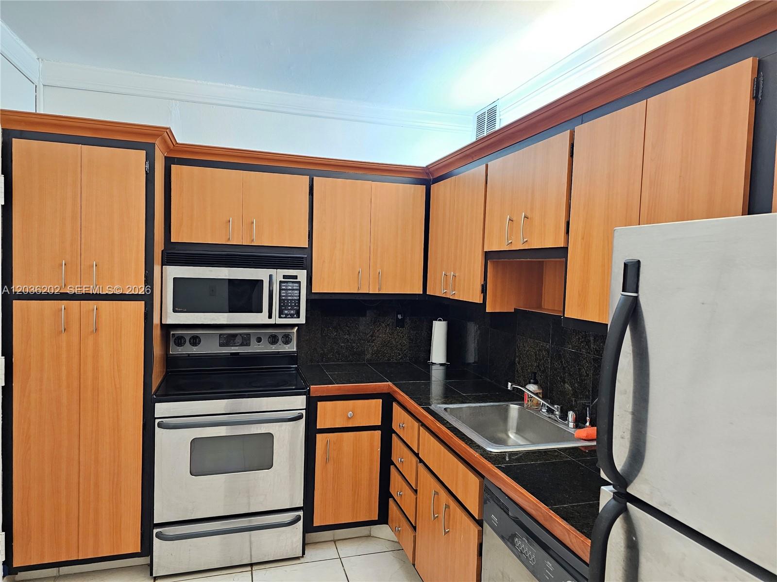 A kitchen features light wood cabinets with black trim, stainless steel appliances, and black countertops — HALLANDALE BEACH, FL