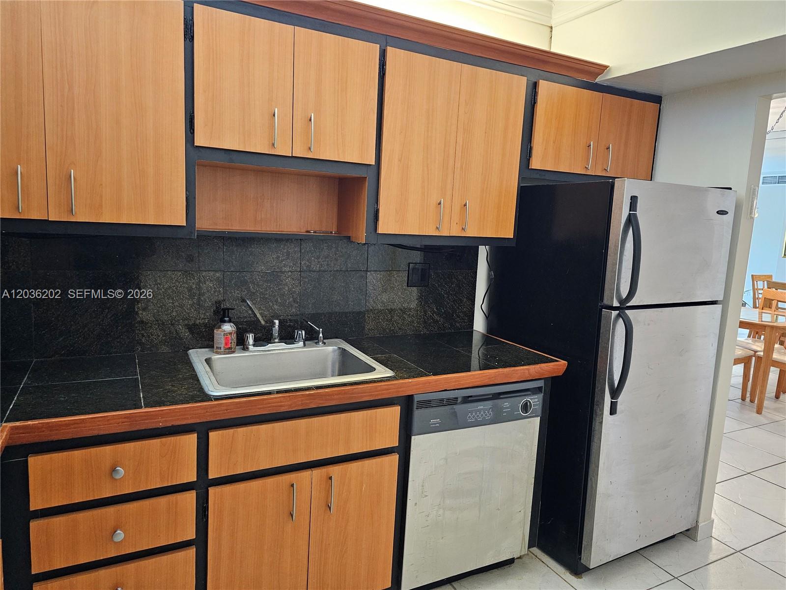A kitchen features wood-tone cabinets, black countertops, a stainless steel sink, and a refrigerator — HALLANDALE BEACH, FL