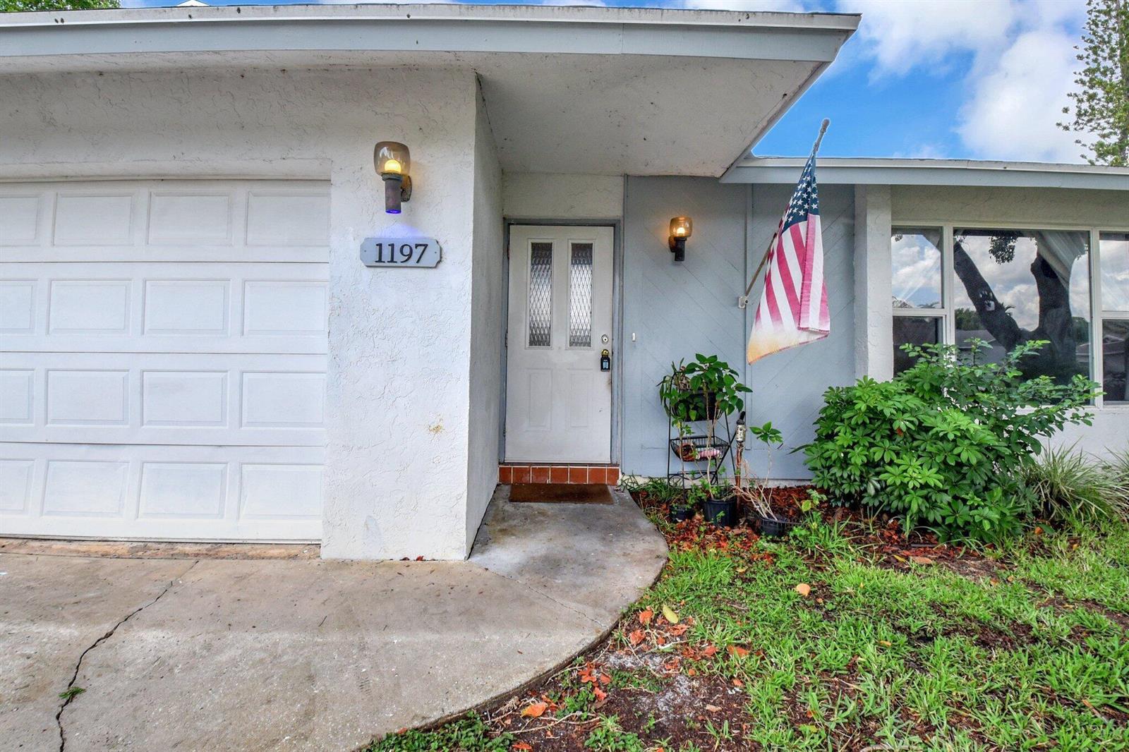 The exterior of a house features a white garage door, a front door, and an American flag — 1197 Belmore Terrace, Wellington FL 33414, WELLINGTON, FL