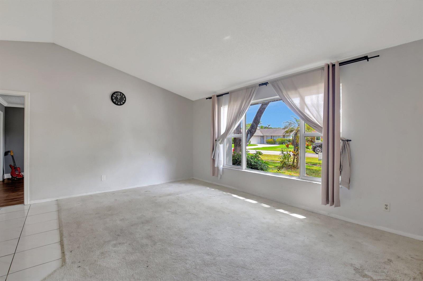 An empty living room features light gray walls, beige carpeting, a large window, and a clock — 1197 Belmore Terrace, Wellington FL 33414, WELLINGTON, FL