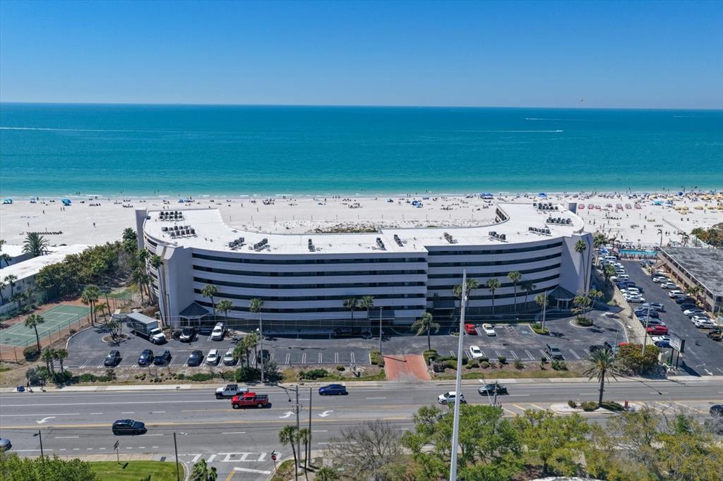 An aerial view shows a curved beachfront hotel building with a parking lot and road — 4450 GULF BOULEVARD UNIT 110, ST PETE BEACH, FL