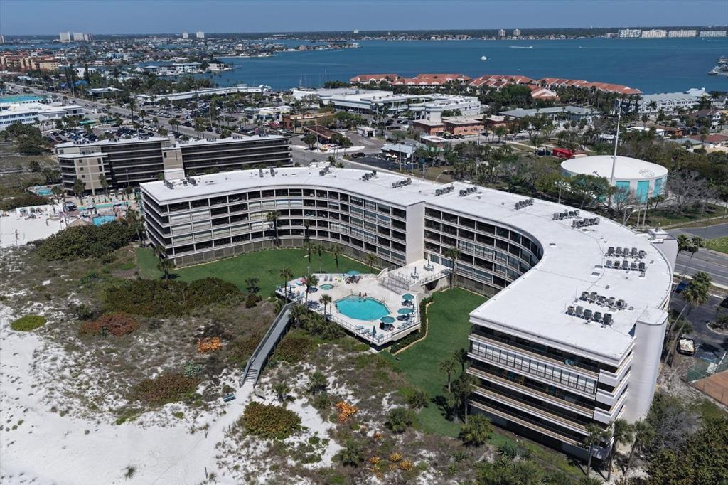 An aerial view of a curved beachfront condominium building with a pool and sandy dunes — 4450 GULF BOULEVARD UNIT 110, ST PETE BEACH, FL