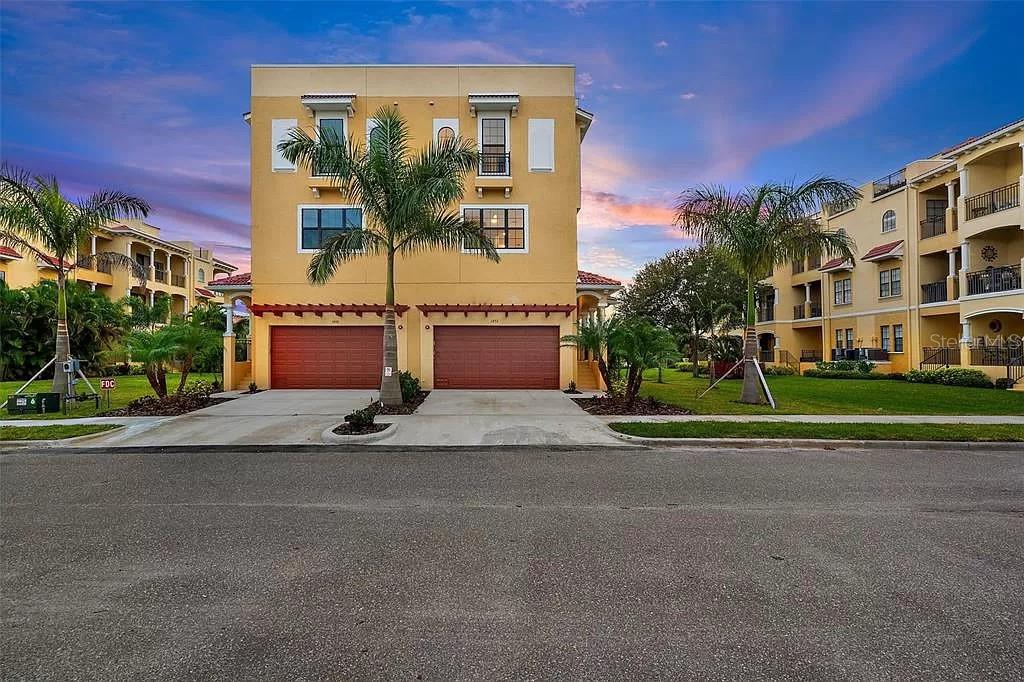 A three-story yellow residential building with two garage doors and palm trees in front — 6418 MARGARITA SHORES LANE, APOLLO BEACH, FL