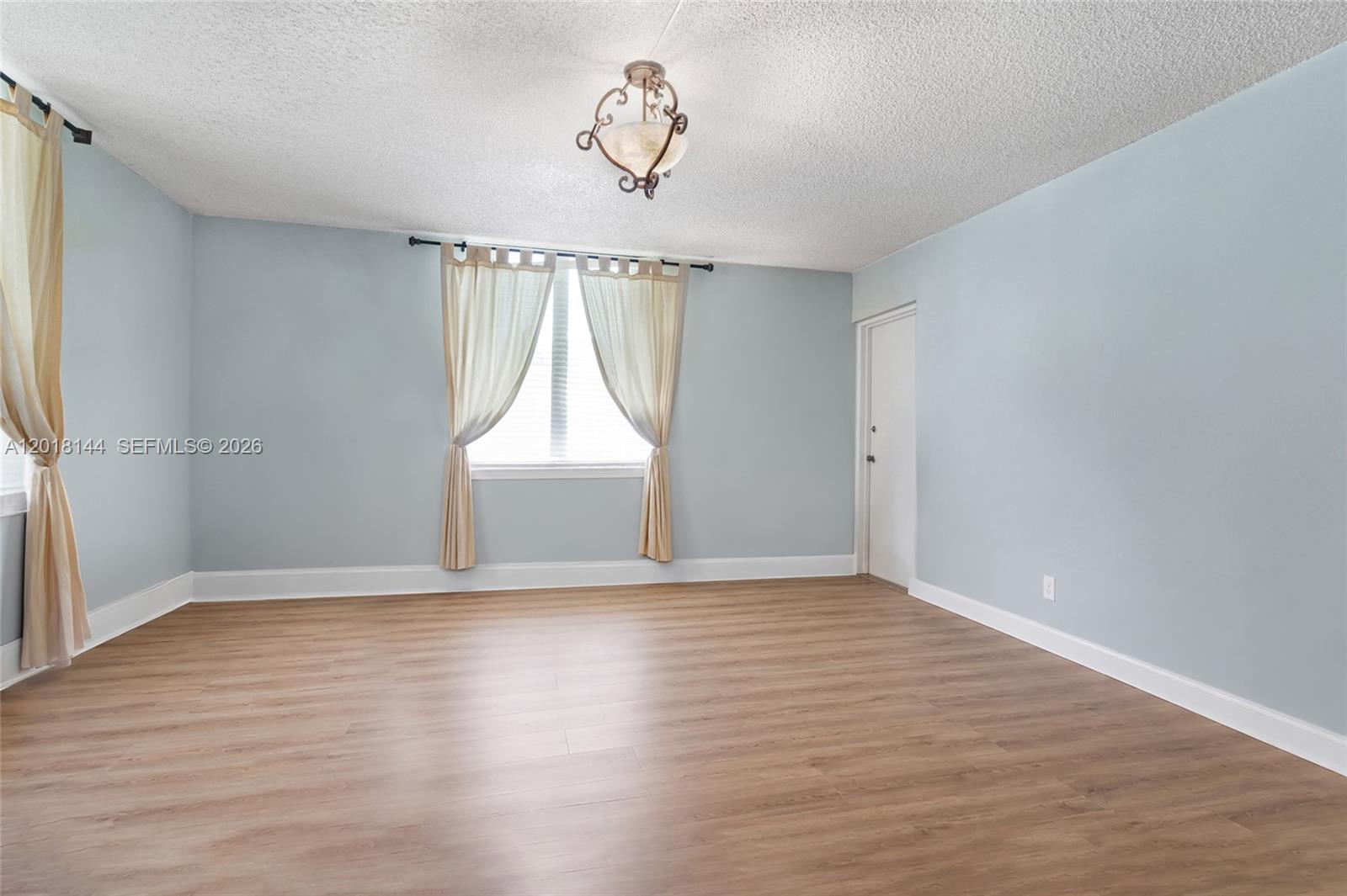 An empty room features light blue walls, wood-look flooring, a window with curtains, and a ceiling light — 6841 SW 147th Ave # 1D, Miami FL 33193, MIAMI, FL