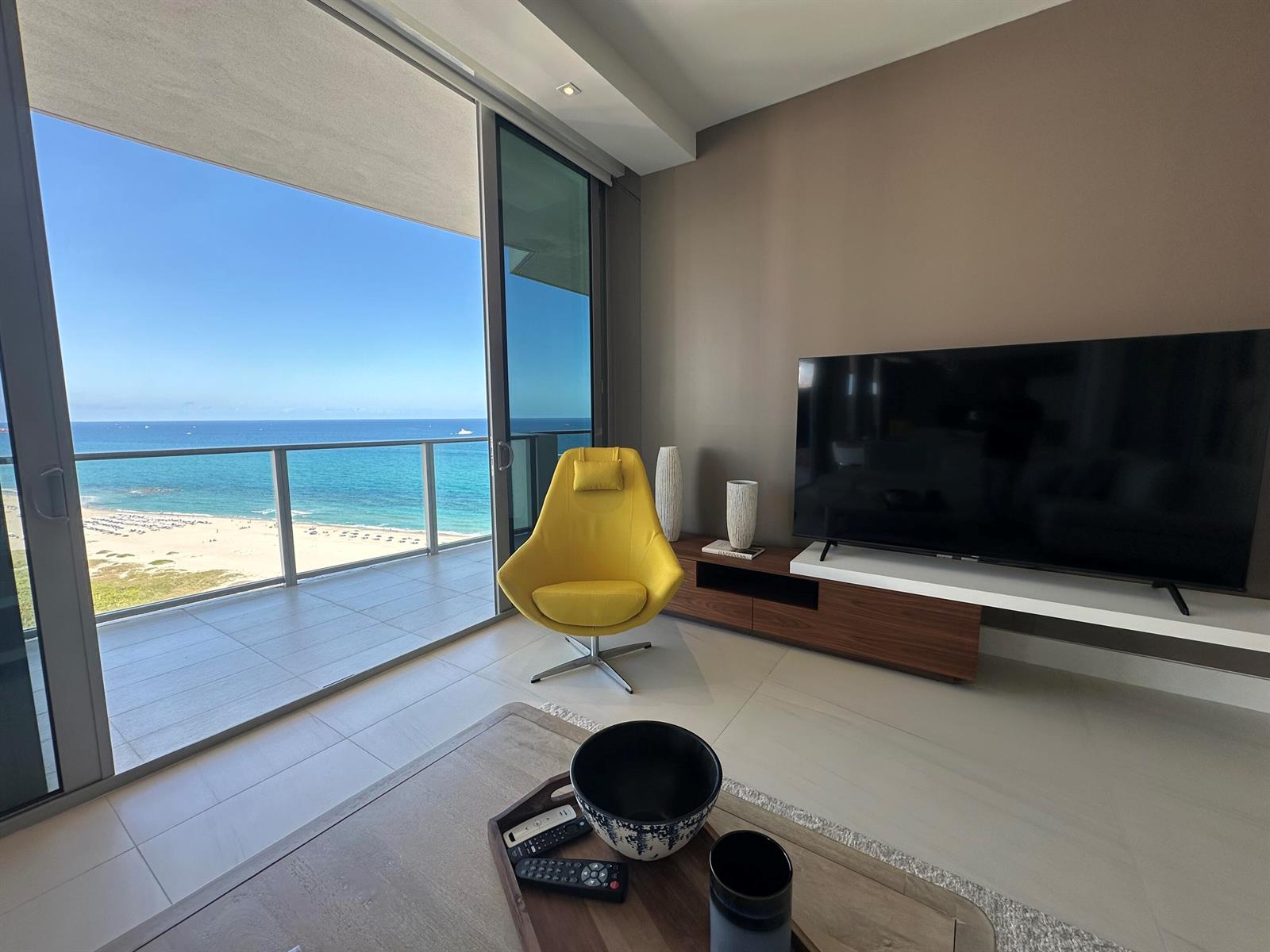 A living room features a yellow armchair, a television, and a balcony overlooking the ocean — 3100 N Ocean Drive # 1701, Riviera Beach FL 33404, RIVIERA BEACH, FL