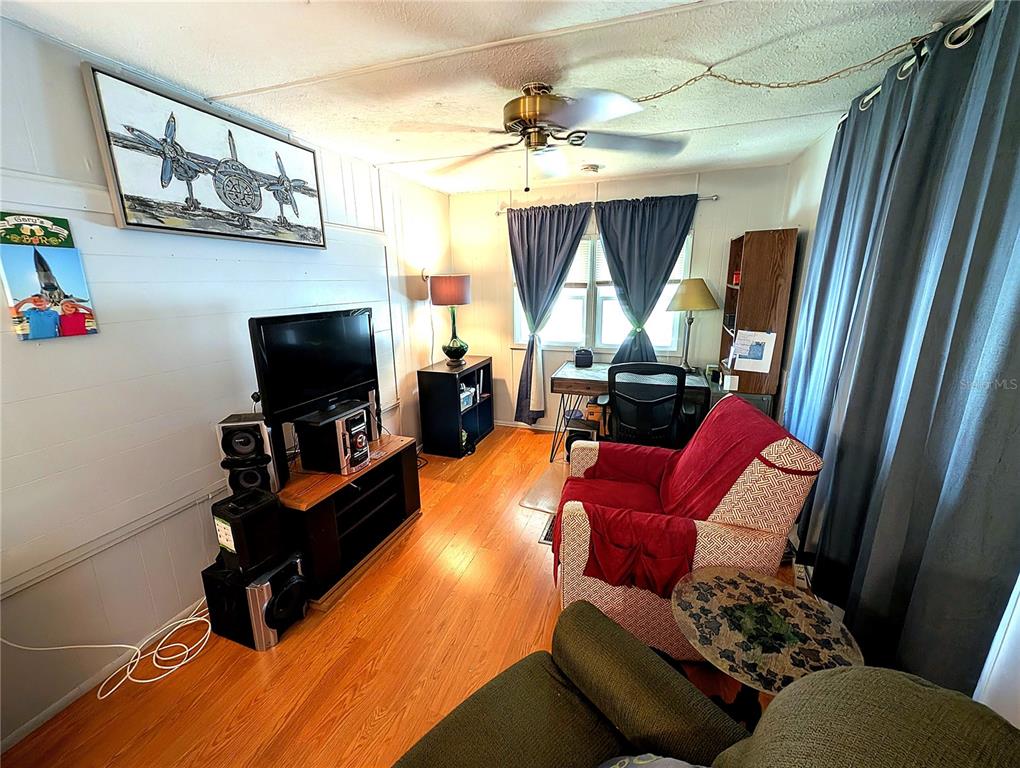 A living room features wood-look flooring, a ceiling fan, a television, and a desk area — 7803 46TH AVENUE N UNIT 49, ST PETERSBURG, FL