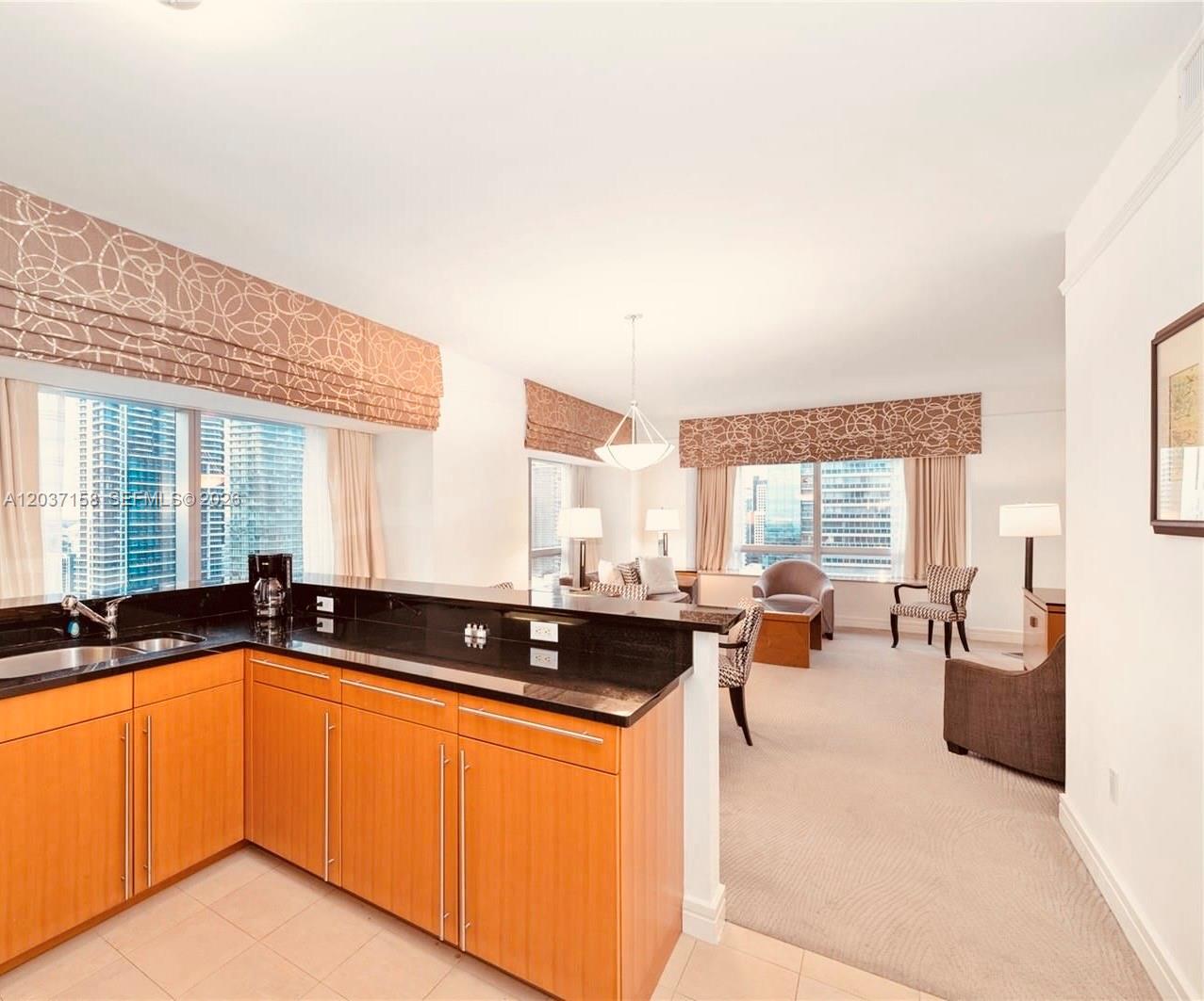 A kitchen with wood cabinets and black countertops overlooks a carpeted living area with windows — 1435 Brickell Ave # 3412, Miami FL 33131, MIAMI, FL