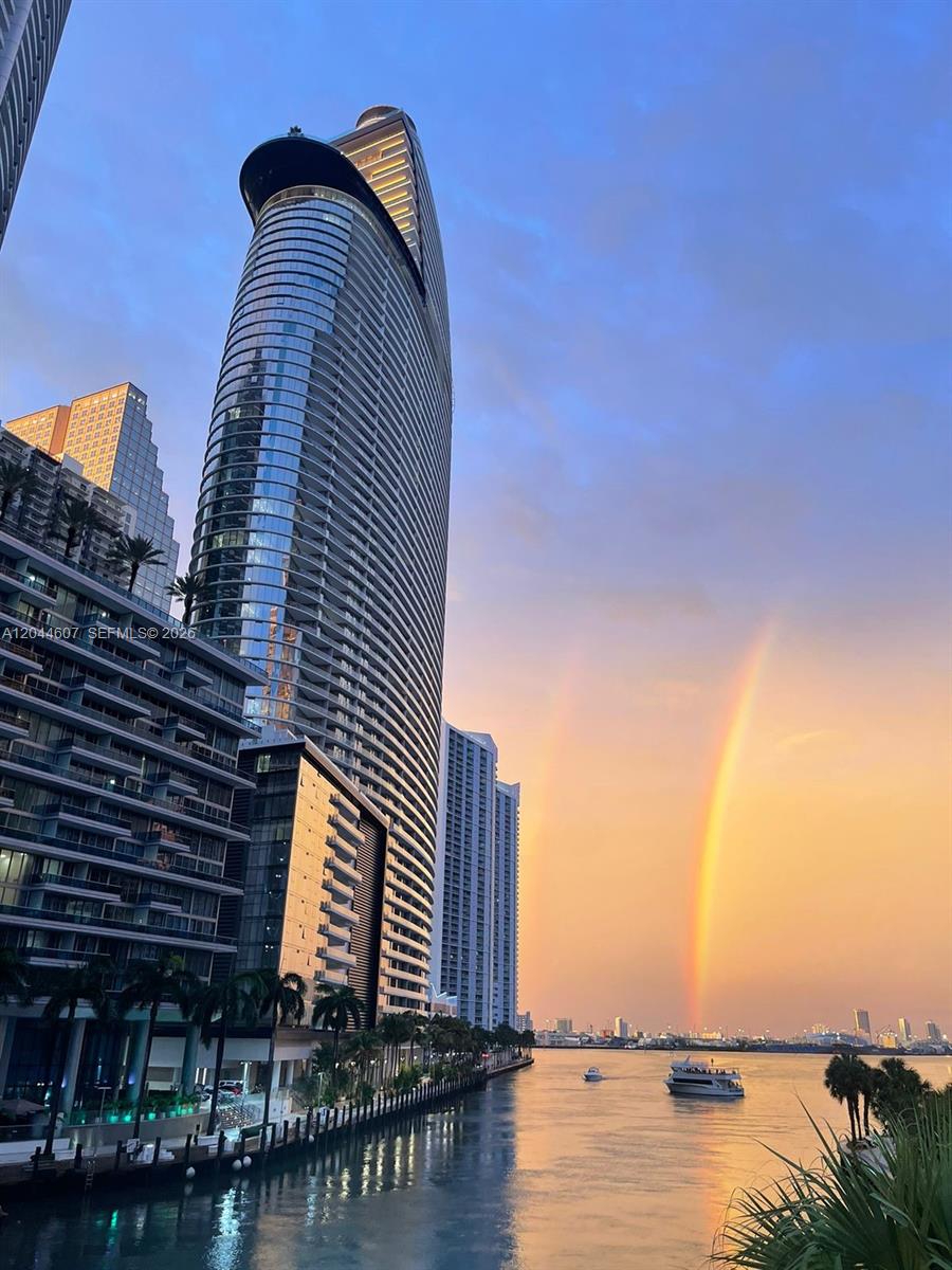 A tall modern skyscraper stands beside a canal under a sunset sky with a rainbow — 300 Biscayne Blvd Way # 1909, Miami FL 33131, MIAMI, FL