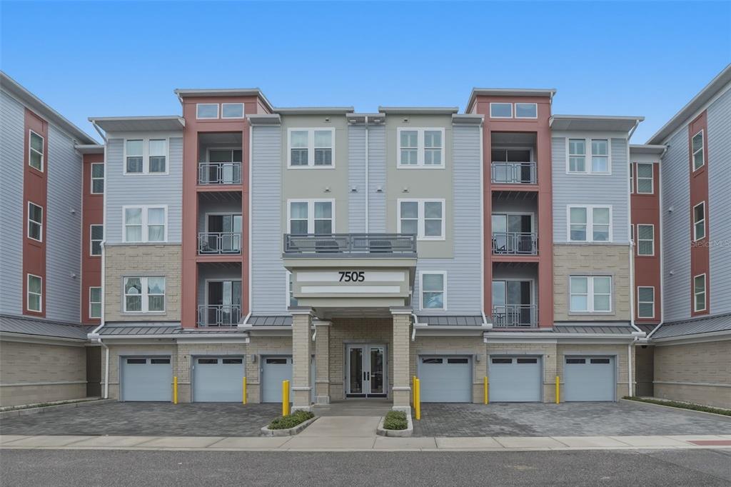 The exterior of a multi-story apartment building features a central entrance, balconies, and ground-level garages — 7505 LAUREATE BOULEVARD UNIT 2408, ORLANDO, FL