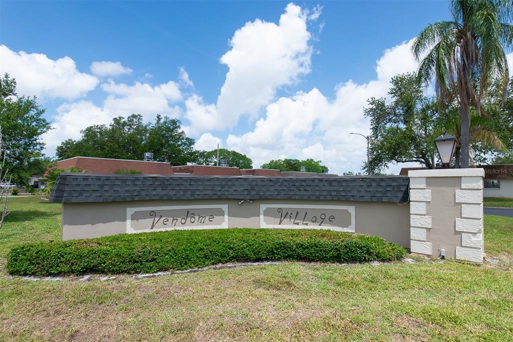 A beige monument sign for Vendome Village sits in a grassy area under a blue sky — 6915 VERSAILLES N, PINELLAS PARK, FL