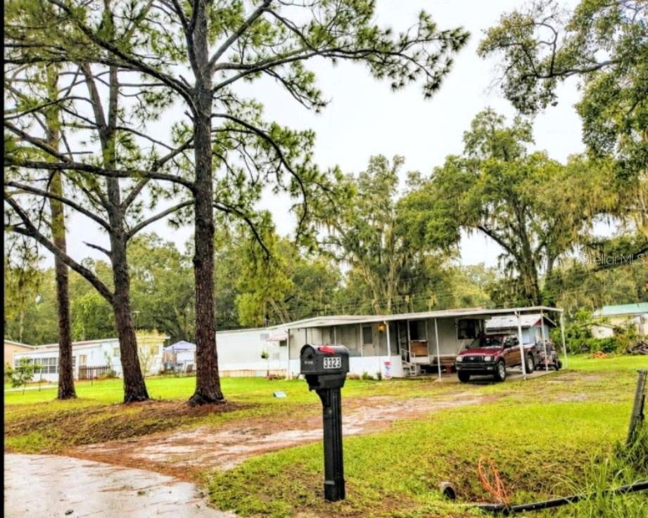 A mobile home with a metal carport sits on a grassy lot with tall trees — 3323 J AND J MANOR ROAD, LAKELAND, FL