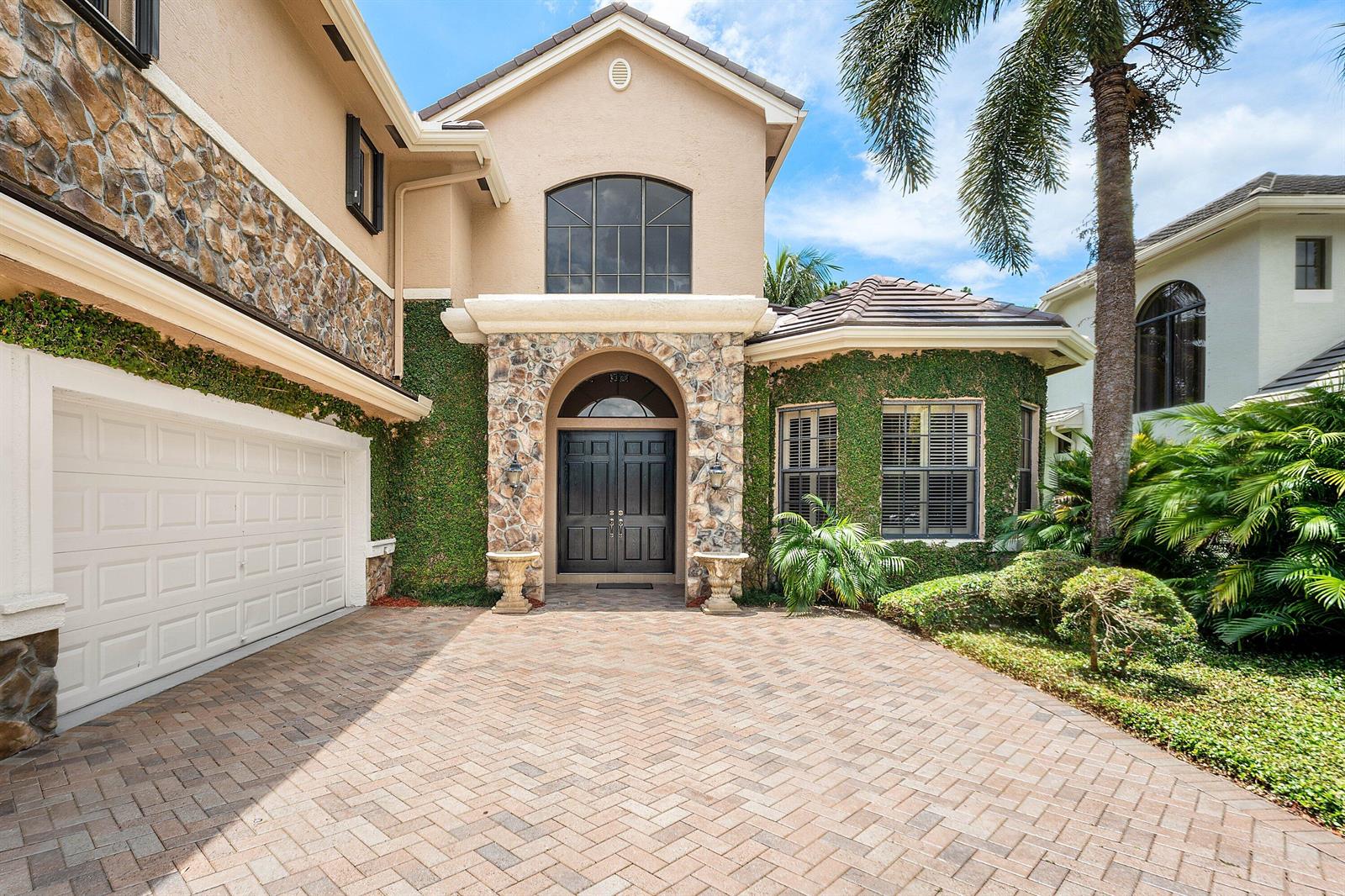 A two-story house exterior features a stone facade, ivy-covered walls, and a paved driveway — 10380 Trianon Place, Wellington FL 33449, WELLINGTON, FL