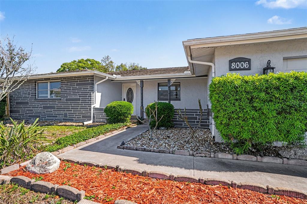 A single-story house features a gray stone facade, a white front door, and landscaped yard — 8006 MIMOSA DRIVE, PORT RICHEY, FL