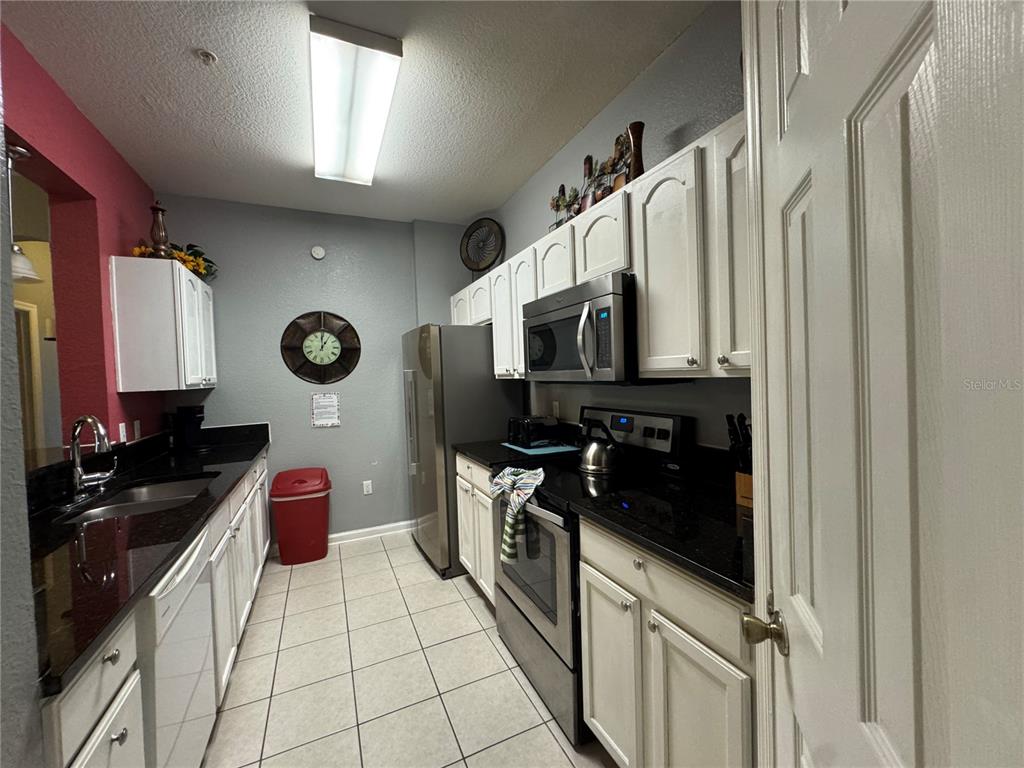 A galley-style kitchen features white cabinets, black countertops, stainless steel appliances, and tiled flooring — 7664 COMROW STREET UNIT 204, KISSIMMEE, FL