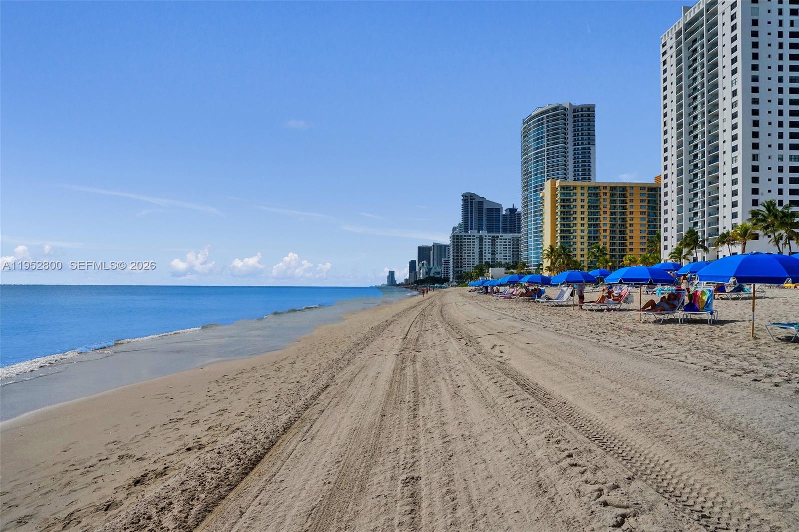 A sandy beach with rows of blue umbrellas and lounge chairs sits before tall buildings — 2201 S Ocean Dr # 608, Hollywood FL 33019, HOLLYWOOD, FL