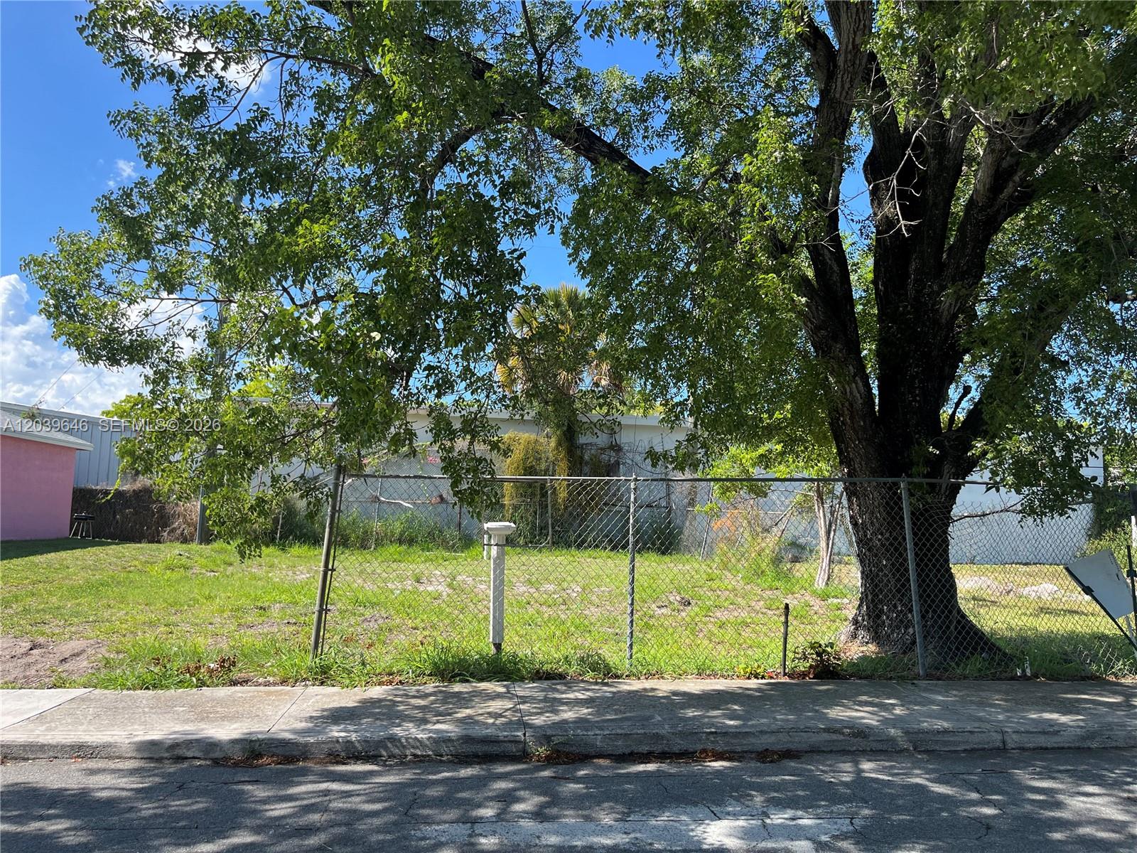 A chain-link fence encloses a grassy lot with a large tree under a blue sky — 535 Lilac CT, West Palm Beach FL 33407, WEST PALM BEACH, FL