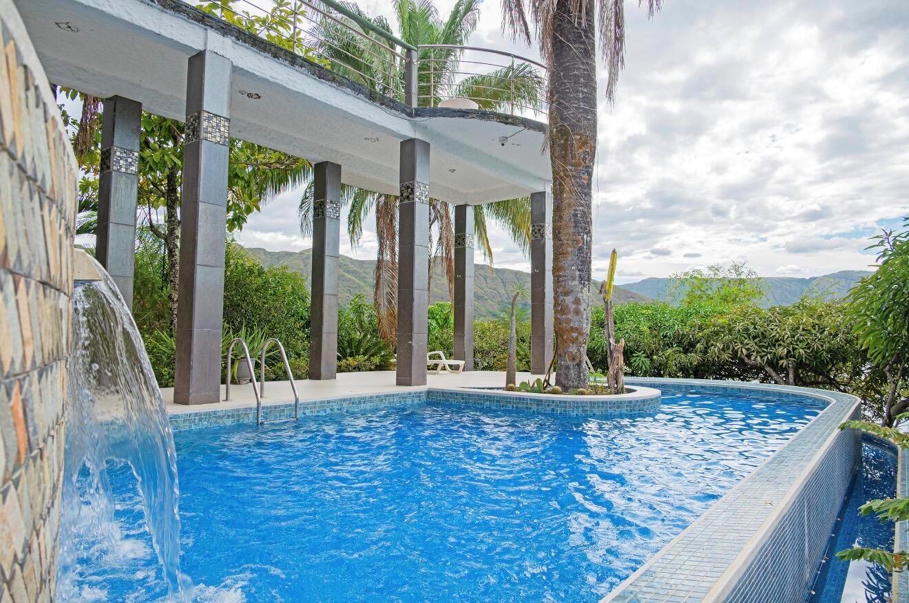 A swimming pool with a waterfall feature sits beneath a covered patio with stone columns — El Paraiso Del Lago, FL