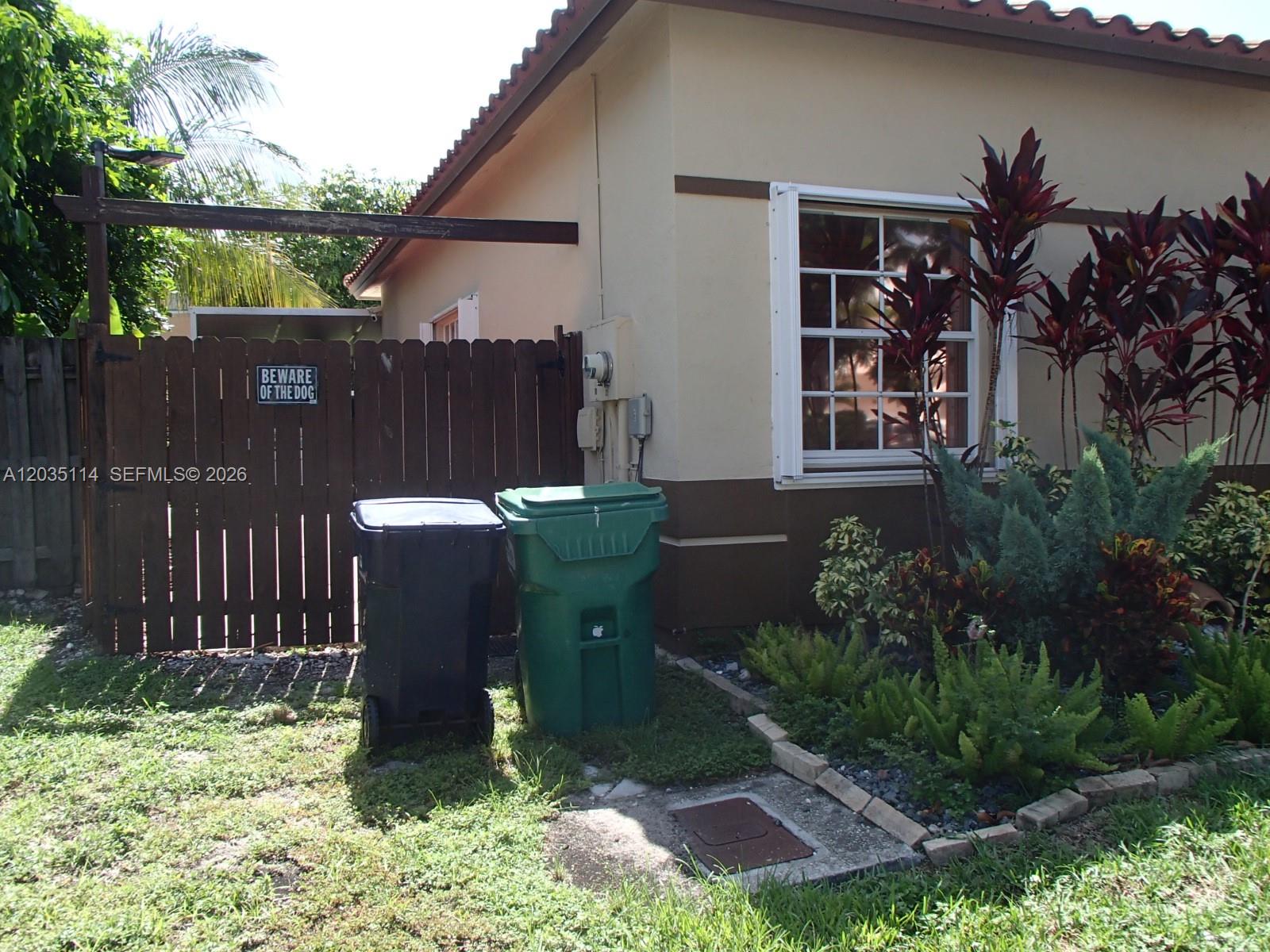 A beige house exterior features a wooden fence, two trash bins, and a landscaped garden — CUTLER BAY, FL