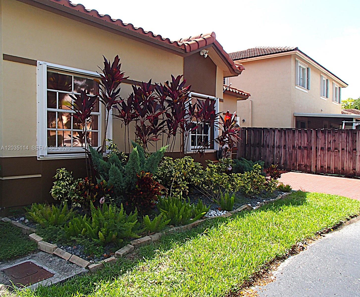 A single-story house exterior features a landscaped garden bed with red-leafed plants and ferns — CUTLER BAY, FL