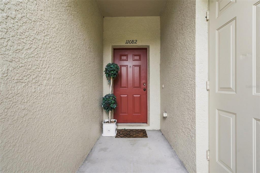 A red front door with a topiary plant sits at the end of a covered entryway — 11082 WINTER CREST DRIVE, RIVERVIEW, FL