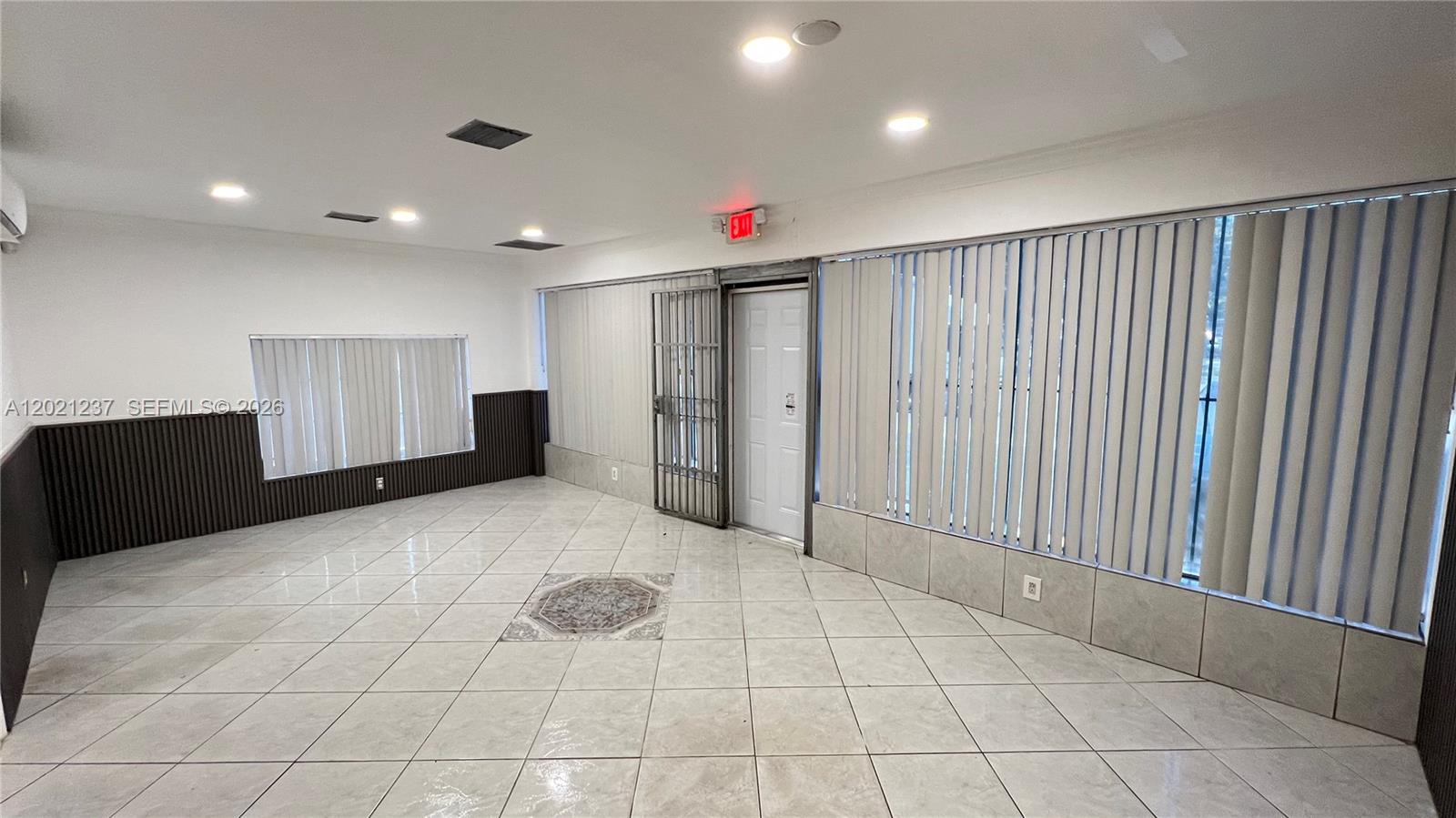 An empty room features white tile flooring, vertical blinds on the windows, and a security gate — 2727 NW 34th St, Miami FL 33142, MIAMI, FL