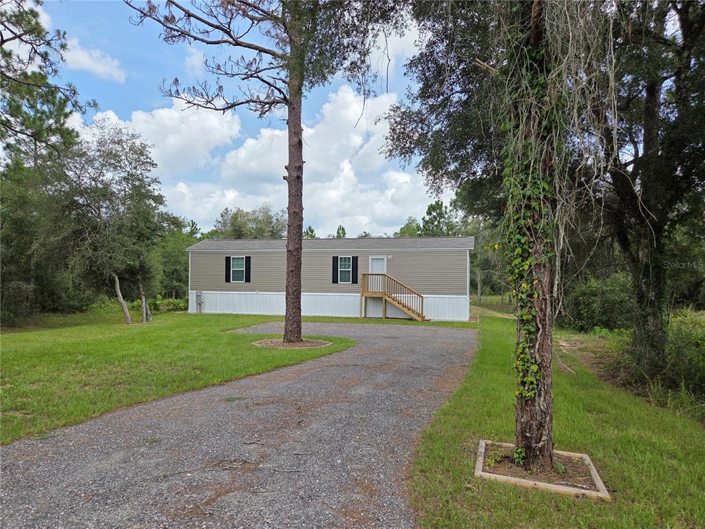 A single-story manufactured home sits on a gravel driveway surrounded by trees and grass — 9448 ROUSSEAU STREET, WEBSTER, FL