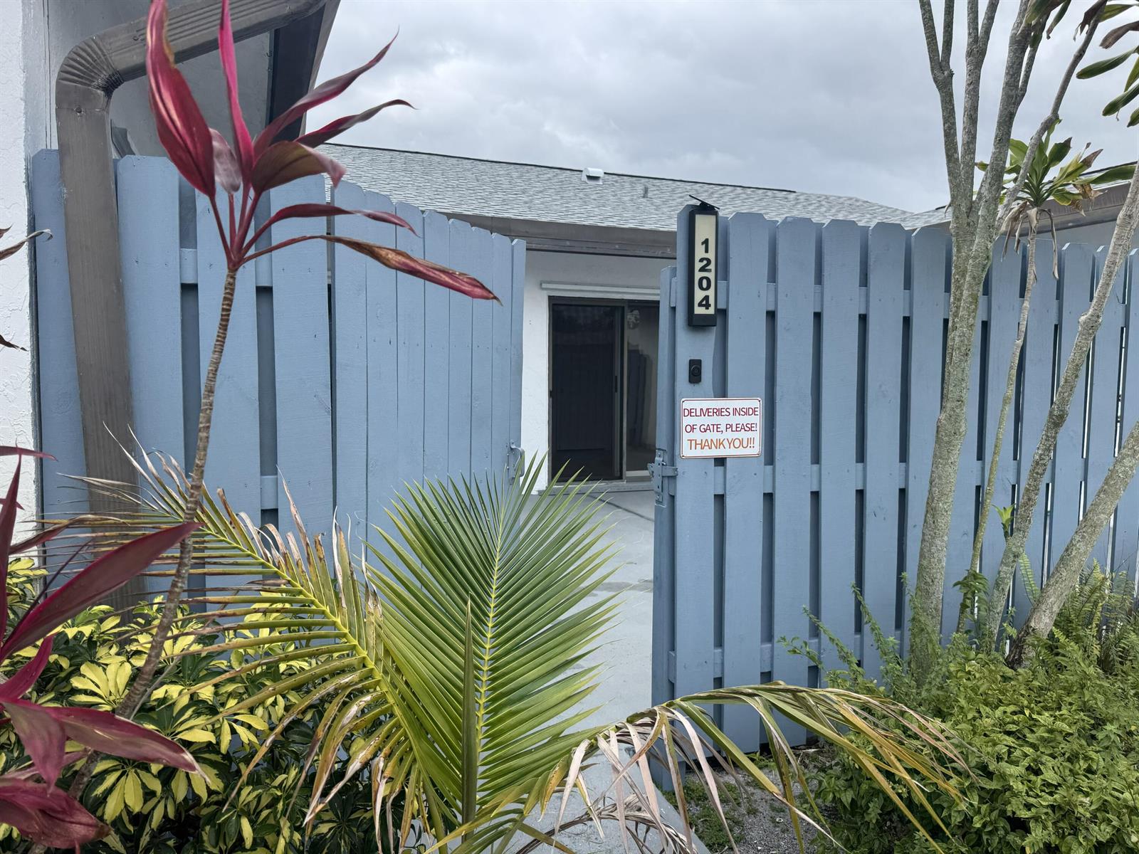 A blue wooden gate with a delivery sign opens to a patio and house entrance — 1204 Timberlane Circle # 12d, FL 33463, FL