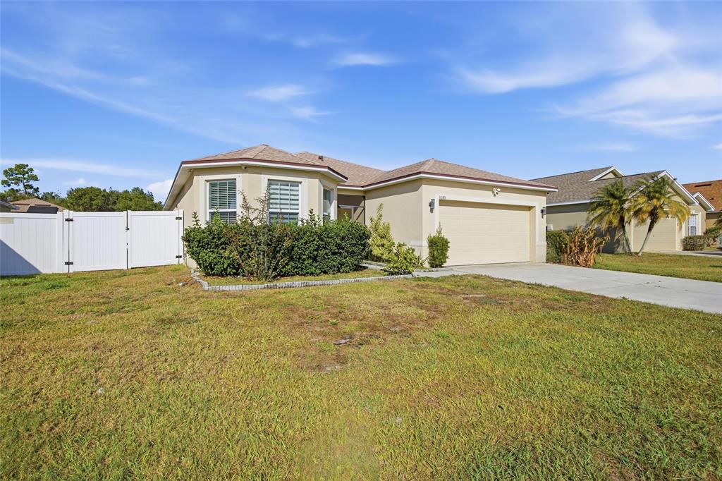 A single-story house with a beige stucco exterior, a two-car garage, and a white fence — 1085 BRENTON MANOR DRIVE, WINTER HAVEN, FL
