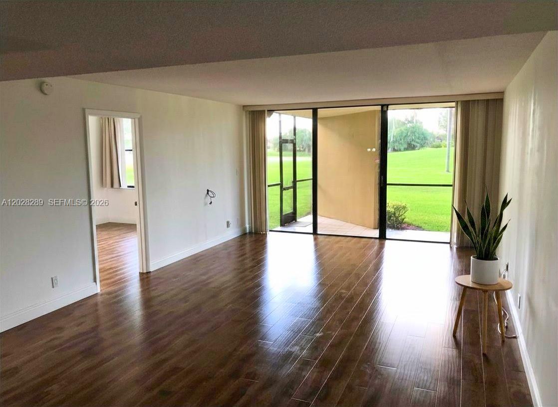 An empty living room features dark wood-look flooring, white walls, and a sliding glass door — 16300 Golf Club Rd # 105, Weston FL 33326, WESTON, FL