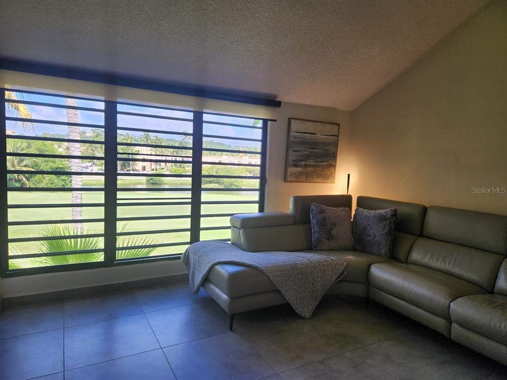 A living room features a grey sectional sofa, tile flooring, and large windows overlooking greenery — 322 Beach Village BEACH VILLAGE - CANDELERO DRIVE - UNIT 322, HUMACAO, PR