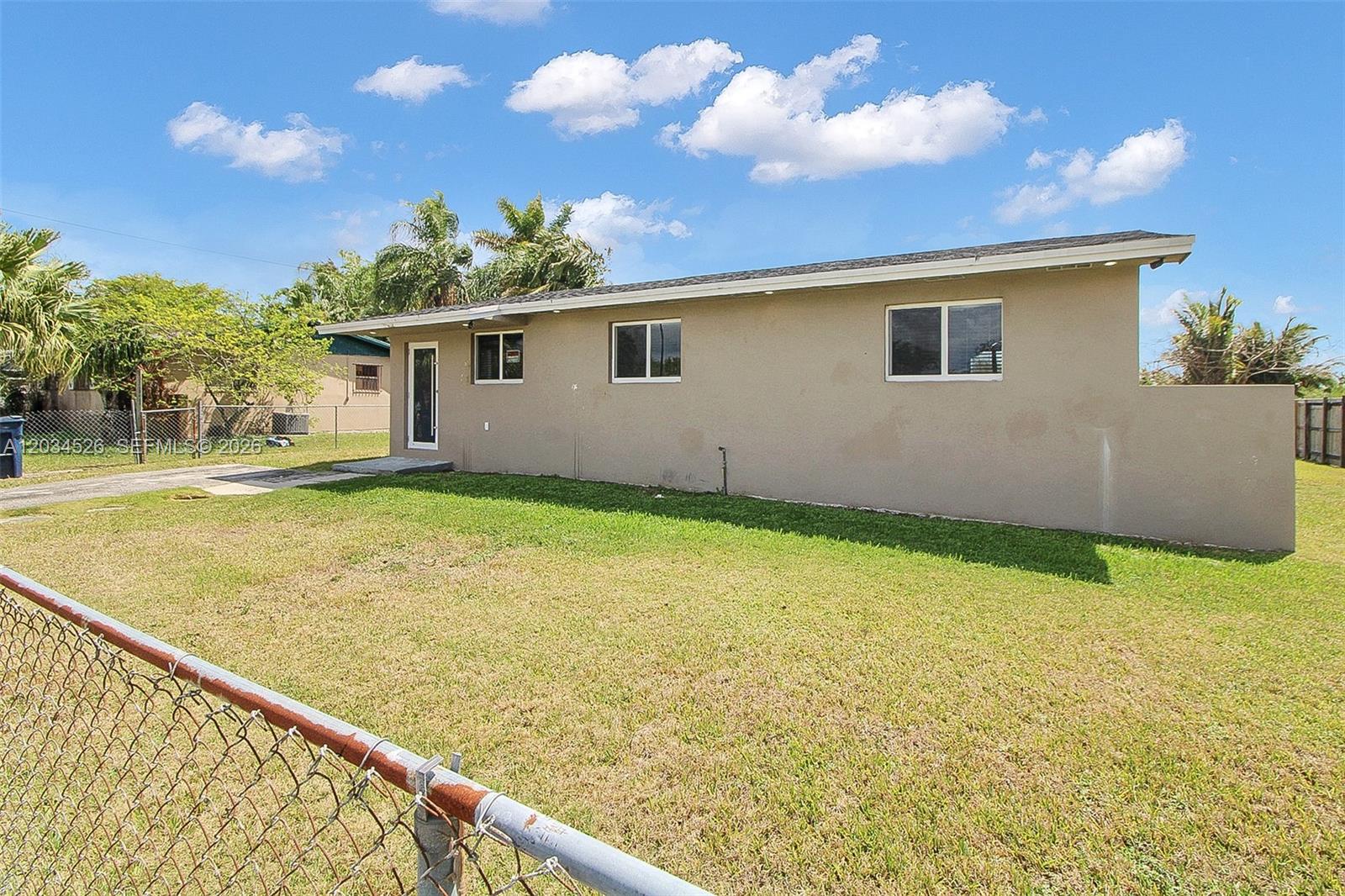 A beige, single-story house exterior features a chain-link fence and a grassy yard — 12840 SW 268th St, Homestead FL 33032, HOMESTEAD, FL