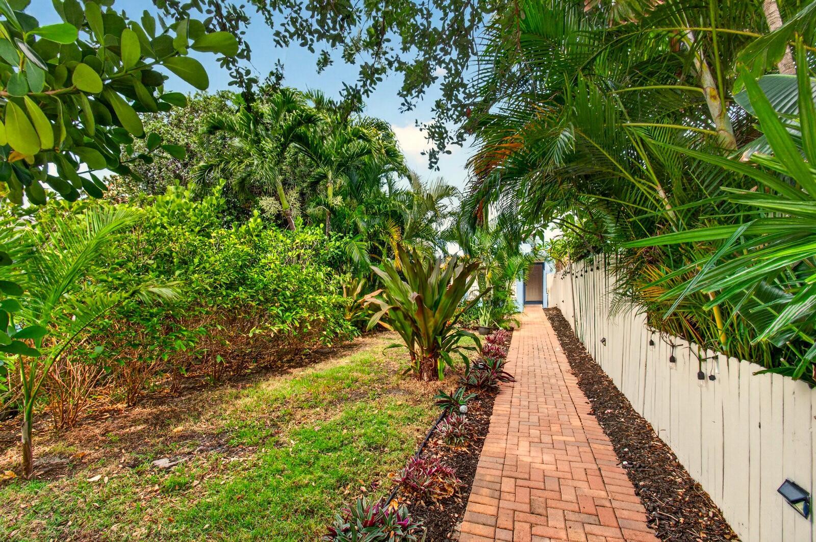 A brick walkway leads through a lush garden toward a blue door beside a fence — 227 N M Street, FL 33460, FL