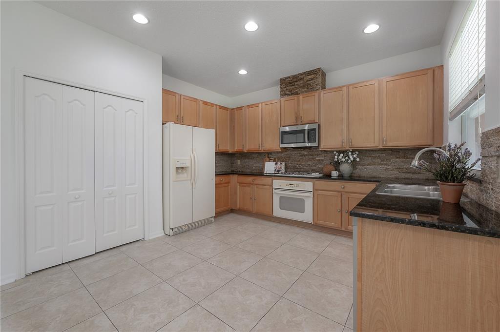 A kitchen features light wood cabinets, dark granite countertops, a stone backsplash, and white appliances — 4716 MAPLETREE LOOP, WESLEY CHAPEL, FL