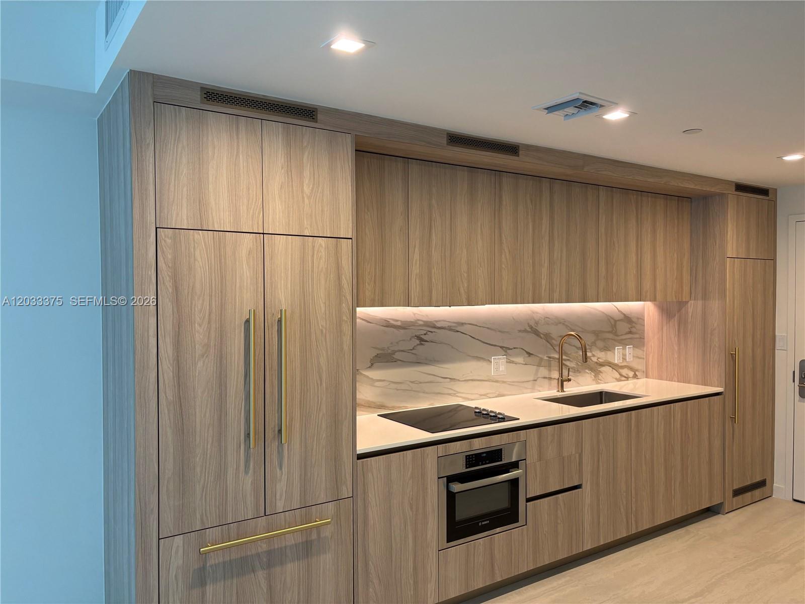 A modern kitchen features wood-grain cabinetry, marble backsplash, gold hardware, and integrated appliances — 77 SE 5th Street # 2108, Miami FL 33131, MIAMI, FL