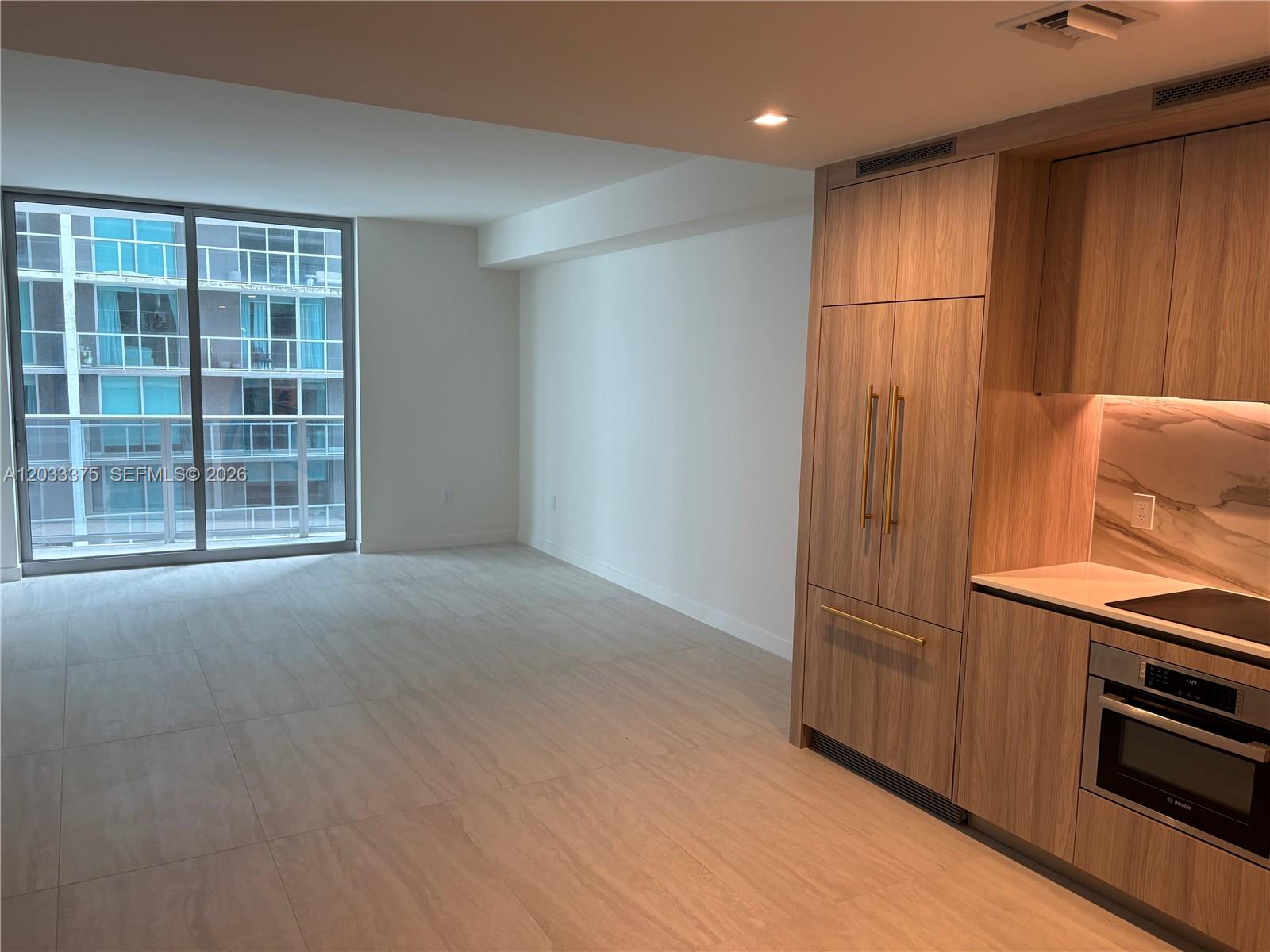A modern kitchen with wood-finish cabinetry and tile flooring opens to an empty living area — 77 SE 5th Street # 2108, Miami FL 33131, MIAMI, FL