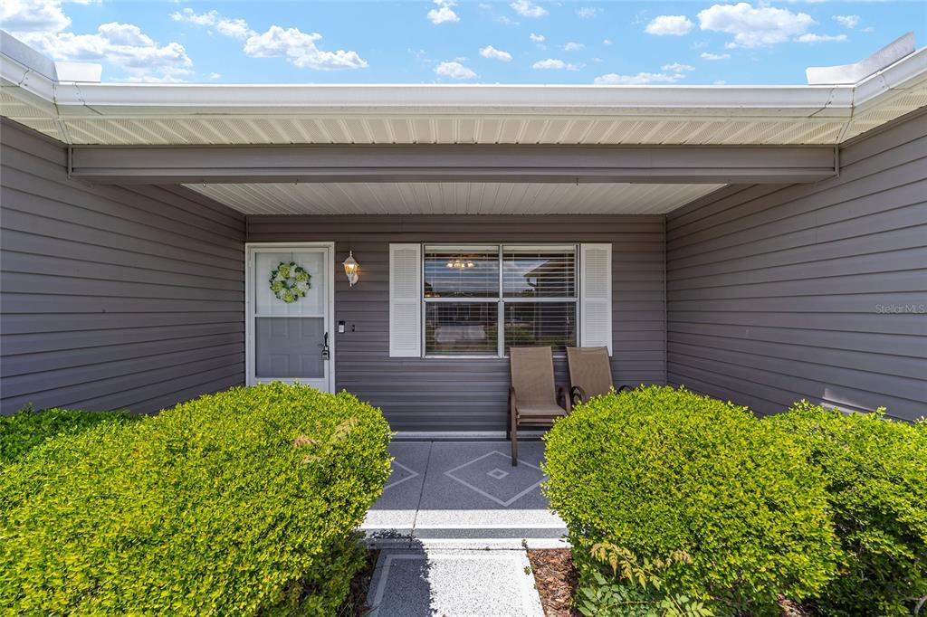 A covered front porch features gray siding, a white door, and two chairs between bushes — 435 SANTA CLARA CIRCLE, THE VILLAGES, FL