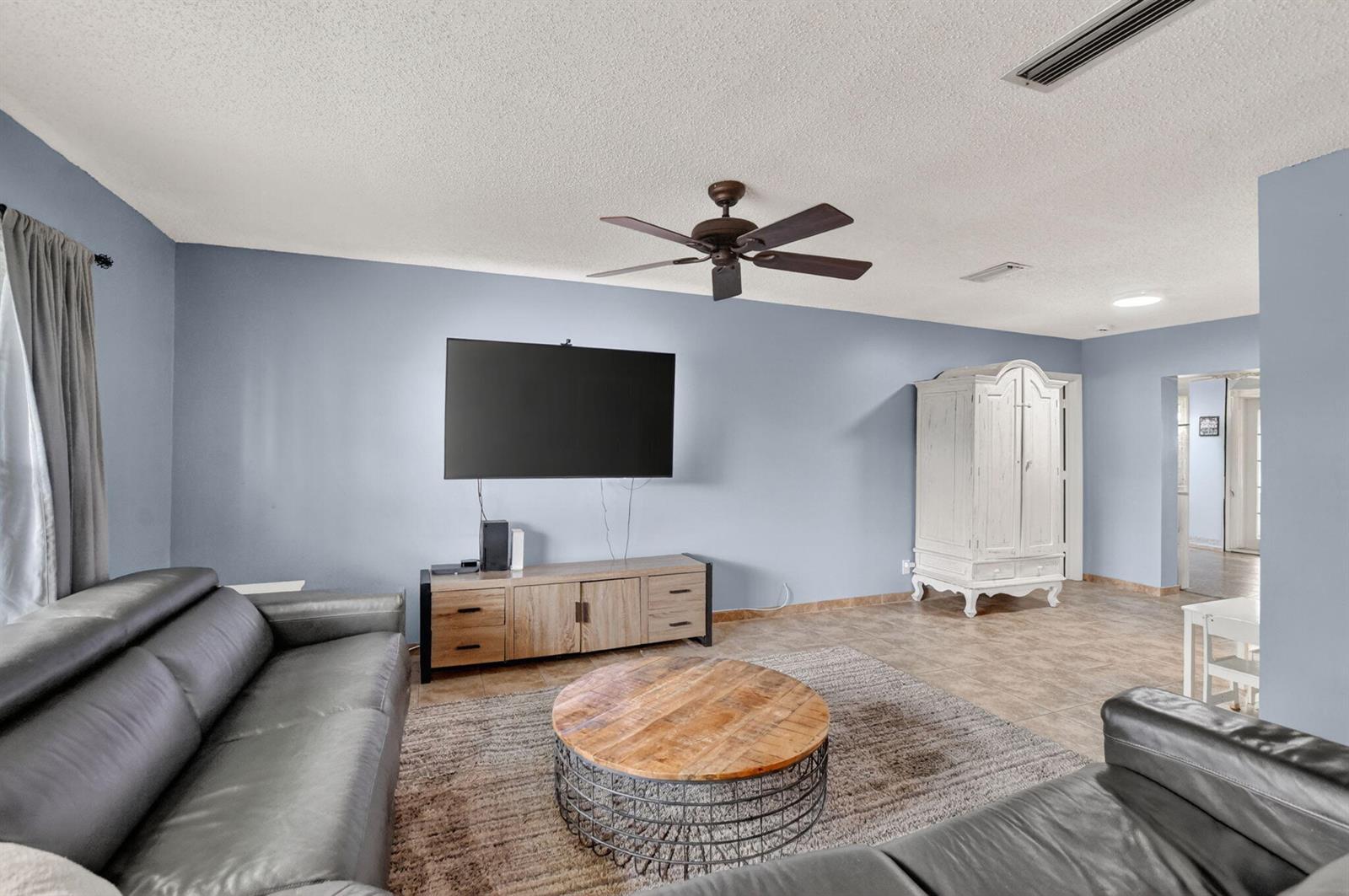 A living room features blue walls, a ceiling fan, a television, and leather sofas — 1140 NW 49th Street, Deerfield Beach FL 33064, DEERFIELD BEACH, FL
